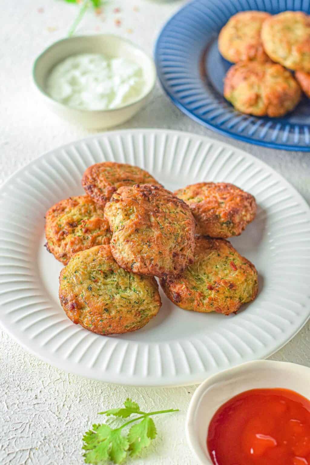 A white plate with several round Air Fryer Zucchini Fritters, accompanied by a bowl of white sauce and a bowl of red sauce on the side. More fritters are on a blue plate in the background.