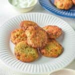 A white plate with several round Air Fryer Zucchini Fritters, accompanied by a bowl of white sauce and a bowl of red sauce on the side. More fritters are on a blue plate in the background.