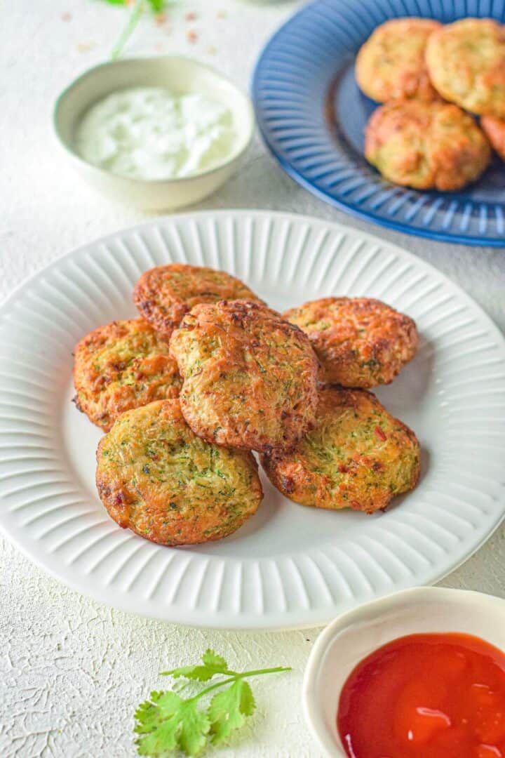 A white plate with several round Air Fryer Zucchini Fritters, accompanied by a bowl of white sauce and a bowl of red sauce on the side. More fritters are on a blue plate in the background.