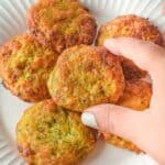 A hand picking up a round, golden-brown cake from a white plate filled with several more.