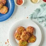 Six Air Fryer Zucchini Fritters are arranged on a white plate, surrounded by small bowls of condiments and a blue plate with more fritters on a textured white surface.