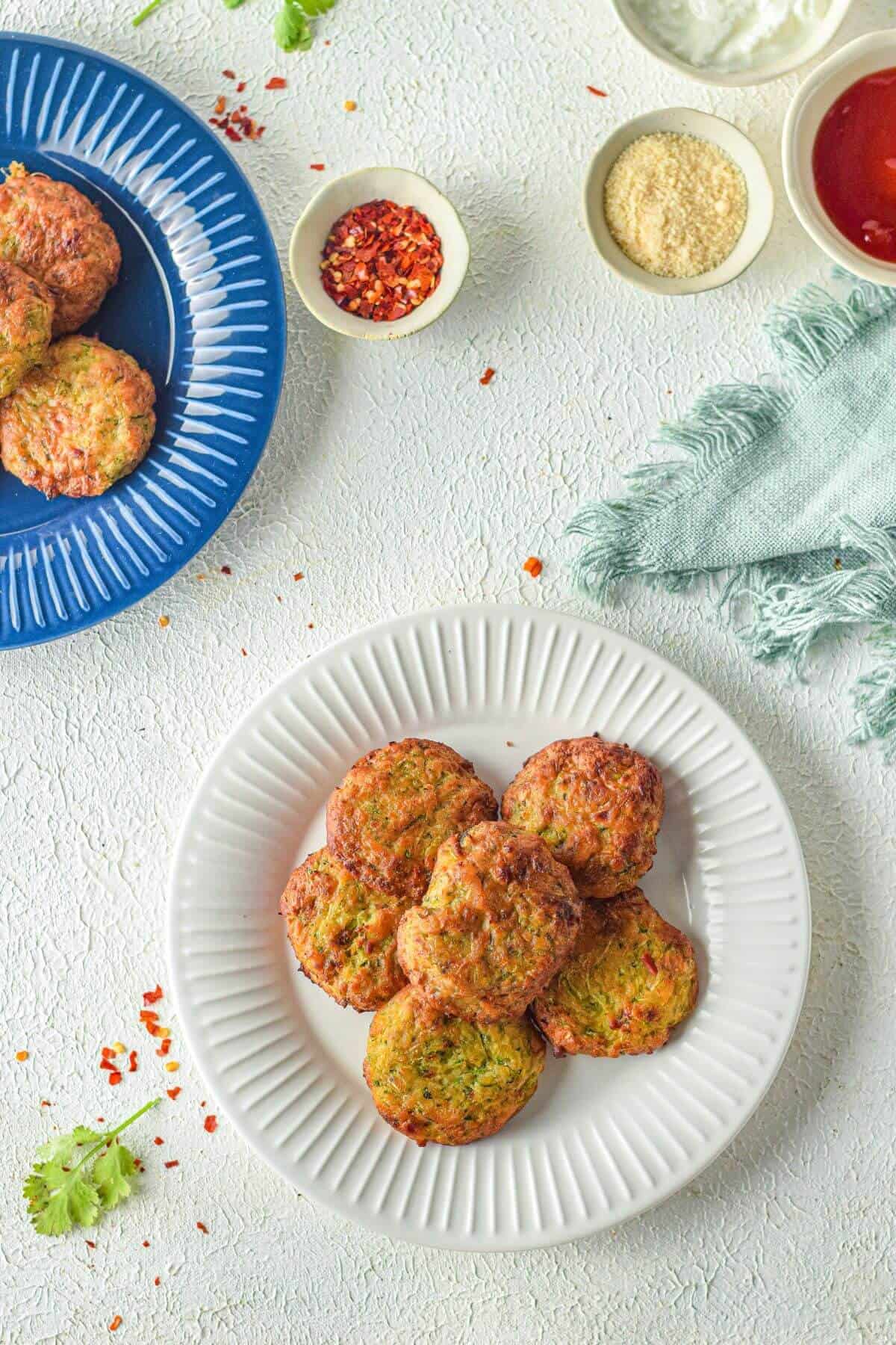 Six cakes are arranged on a white plate, surrounded by small bowls of condiments and a blue plate with more fritters on a textured white surface.