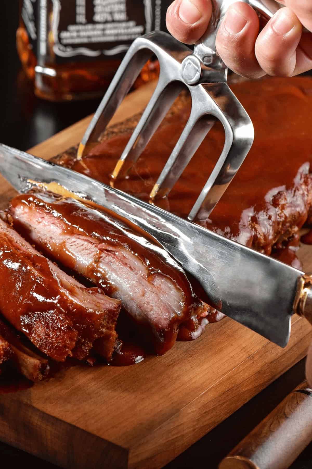 A hand uses a meat fork to hold ribs while slicing through them with a knife on a wooden cutting board. The ribs are coated in a glossy  sauce.