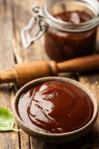 A bowl of bourbon BBQ sauce sits on a wooden surface, with a brush and a jar of the rich, flavorful sauce in the background.