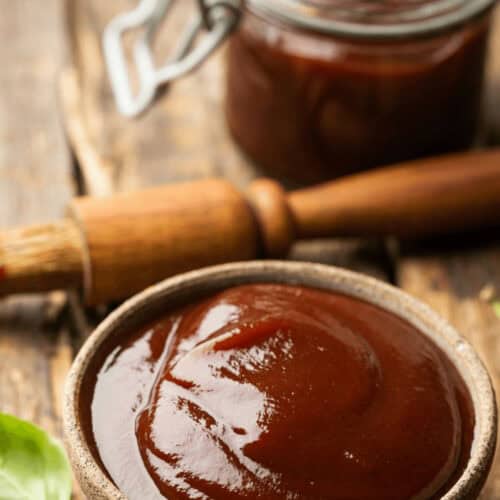 A bowl of bourbon BBQ sauce sits on a wooden surface, with a brush and a jar of the rich, flavorful sauce in the background.