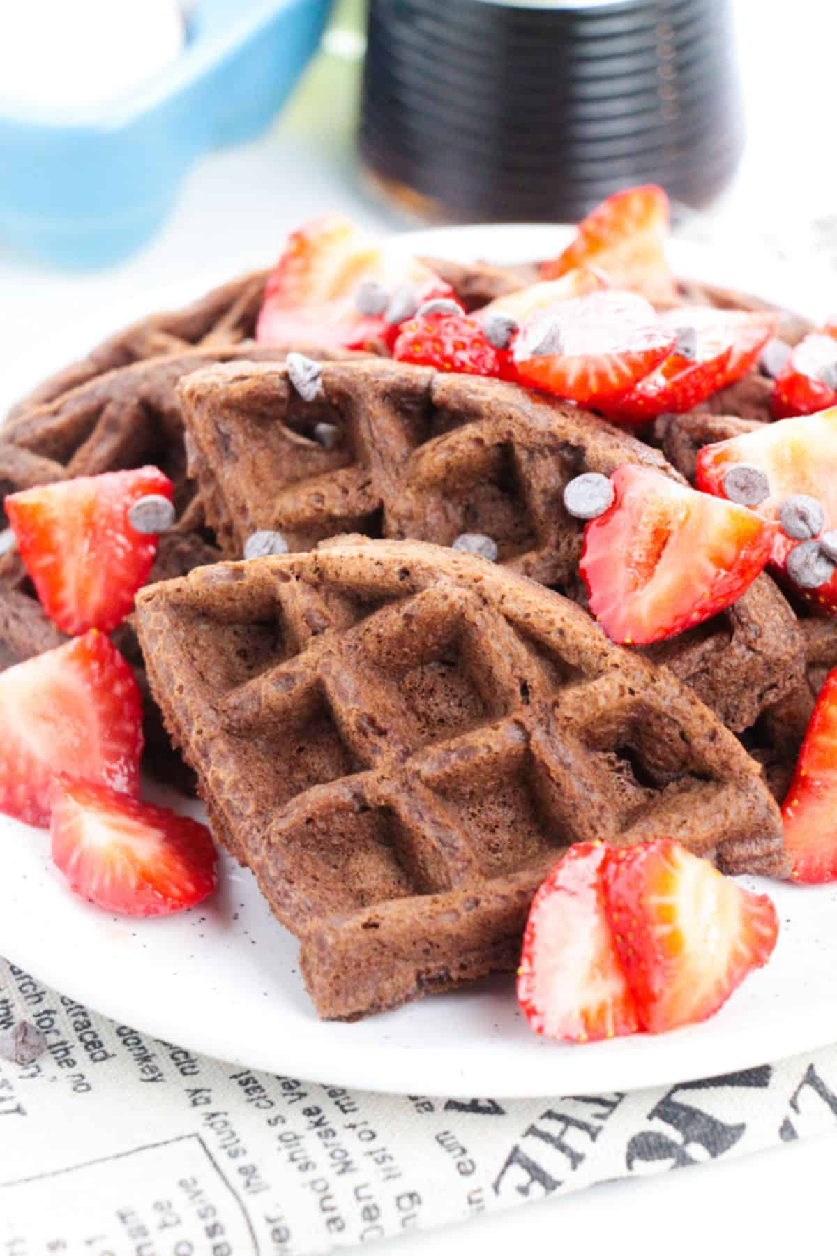Chocolate Waffles topped with sliced strawberries and chocolate chips are artfully arranged on a white plate.