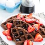 A plate of Chocolate Waffles topped with sliced strawberries and chocolate chips, drizzled with syrup, sits next to a syrup dispenser and a carton of eggs.