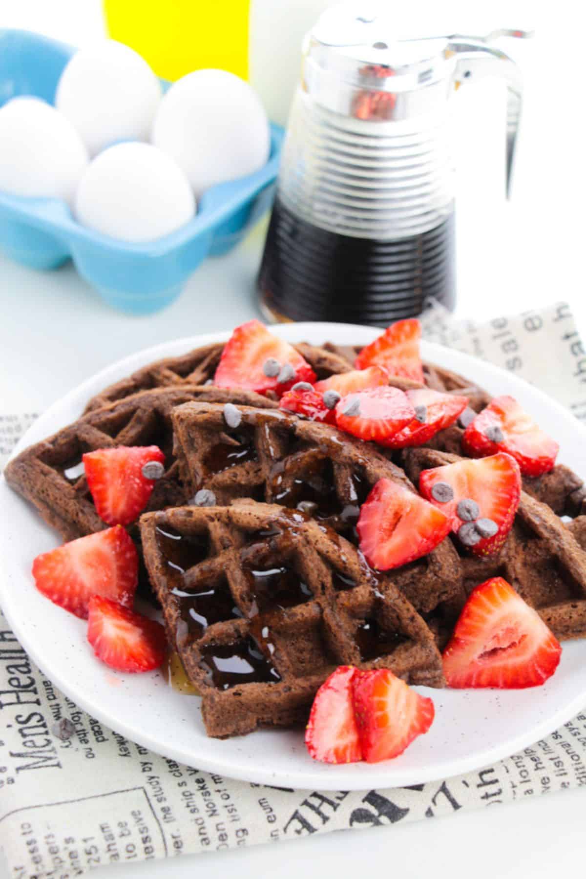 A plate topped with sliced strawberries and chocolate chips, drizzled with syrup, sits next to a syrup dispenser and a carton of eggs.
