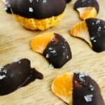 Chocolate Covered Oranges take center stage—tangerine segments and a whole tangerine, partially dipped in dark chocolate, sprinkled with sea salt, and artfully arranged on a wooden surface.