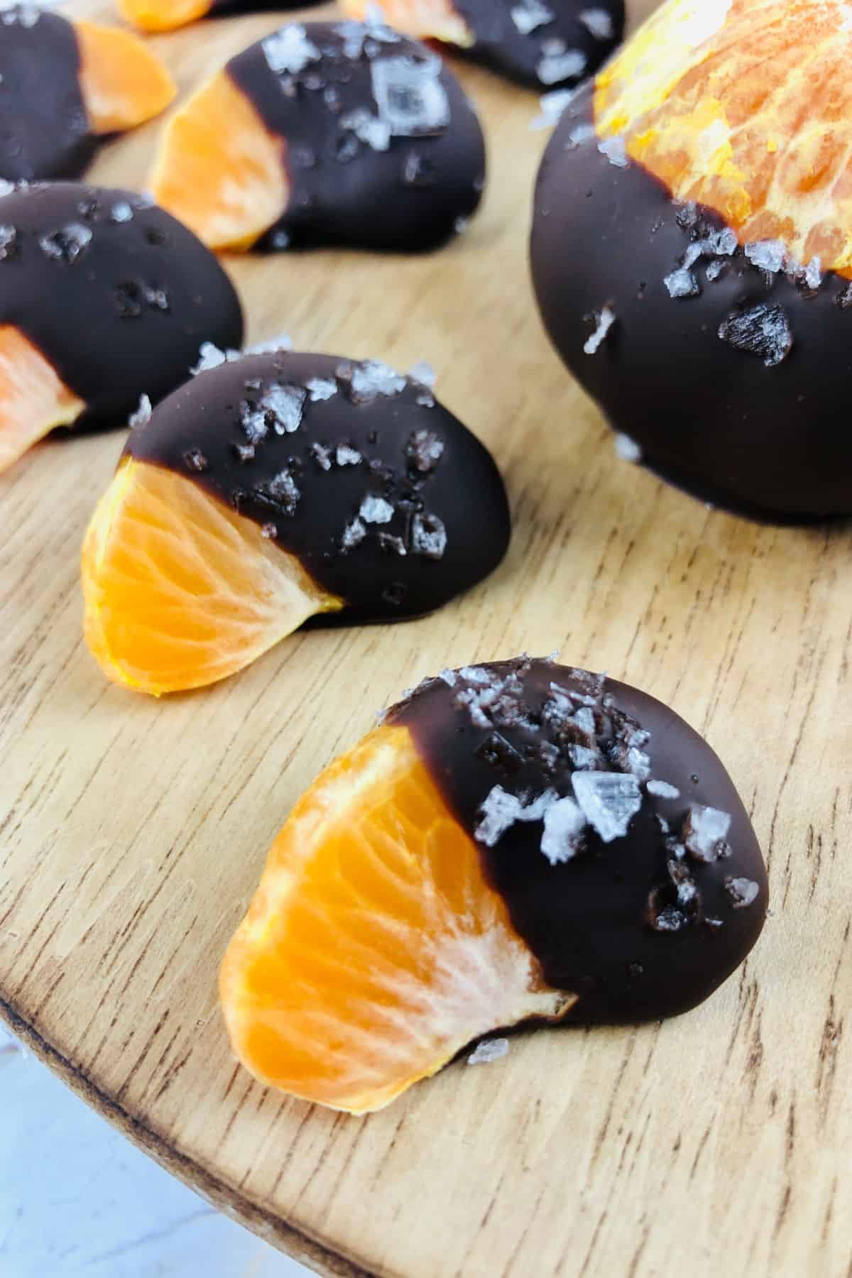 Chocolate Covered Oranges: Orange segments partially dipped in dark chocolate and sprinkled with sea salt, arranged on a wooden surface.