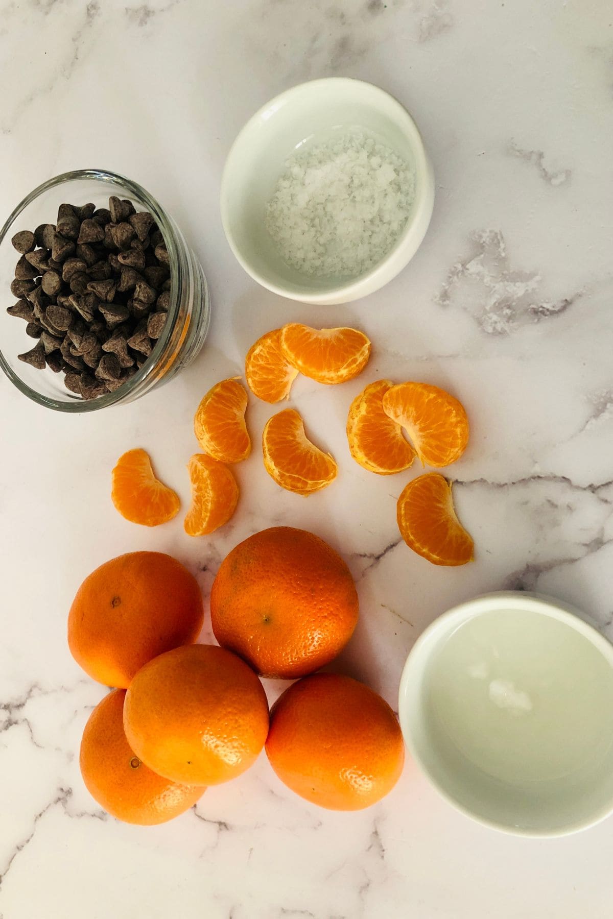 Four whole mandarins, peeled segments, a bowl of chocolate chips, and a bowl of coarse salt are arranged on a white marble surface—perfect for making Chocolate Covered Oranges.