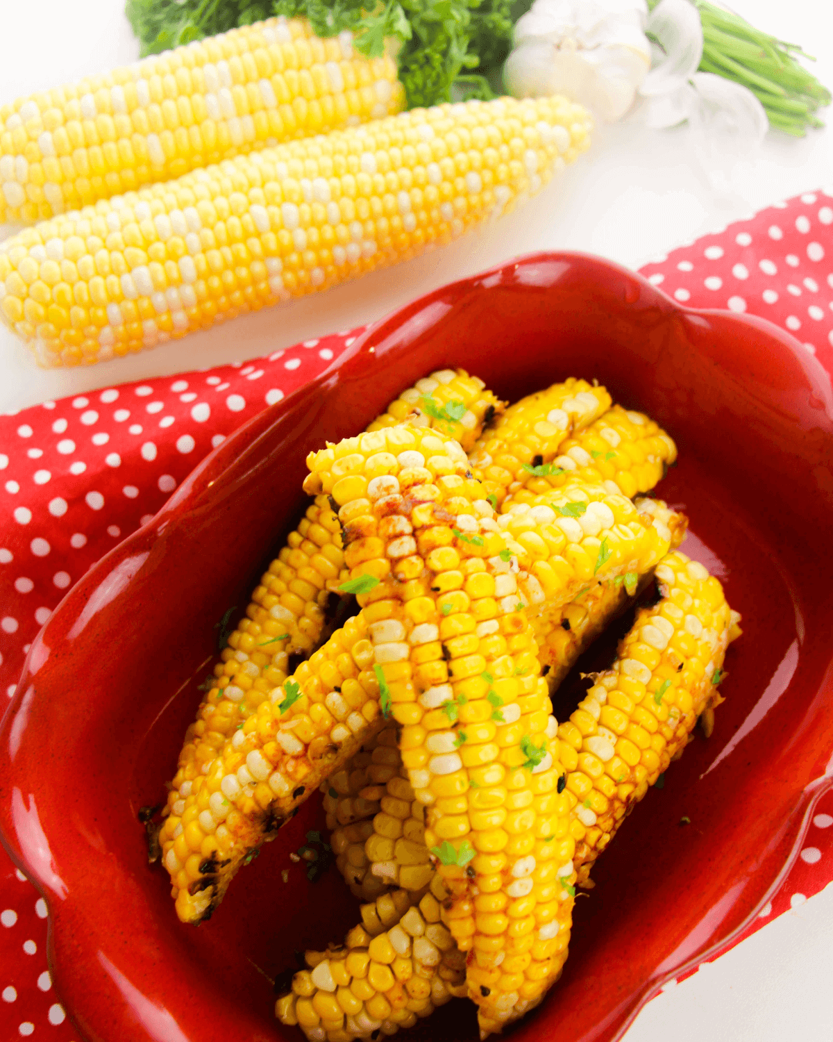 Grilled corn ribs with garlic butter served in a red dish, accompanied by fresh corn and greens in the background, all set on a charming red polka-dot cloth.