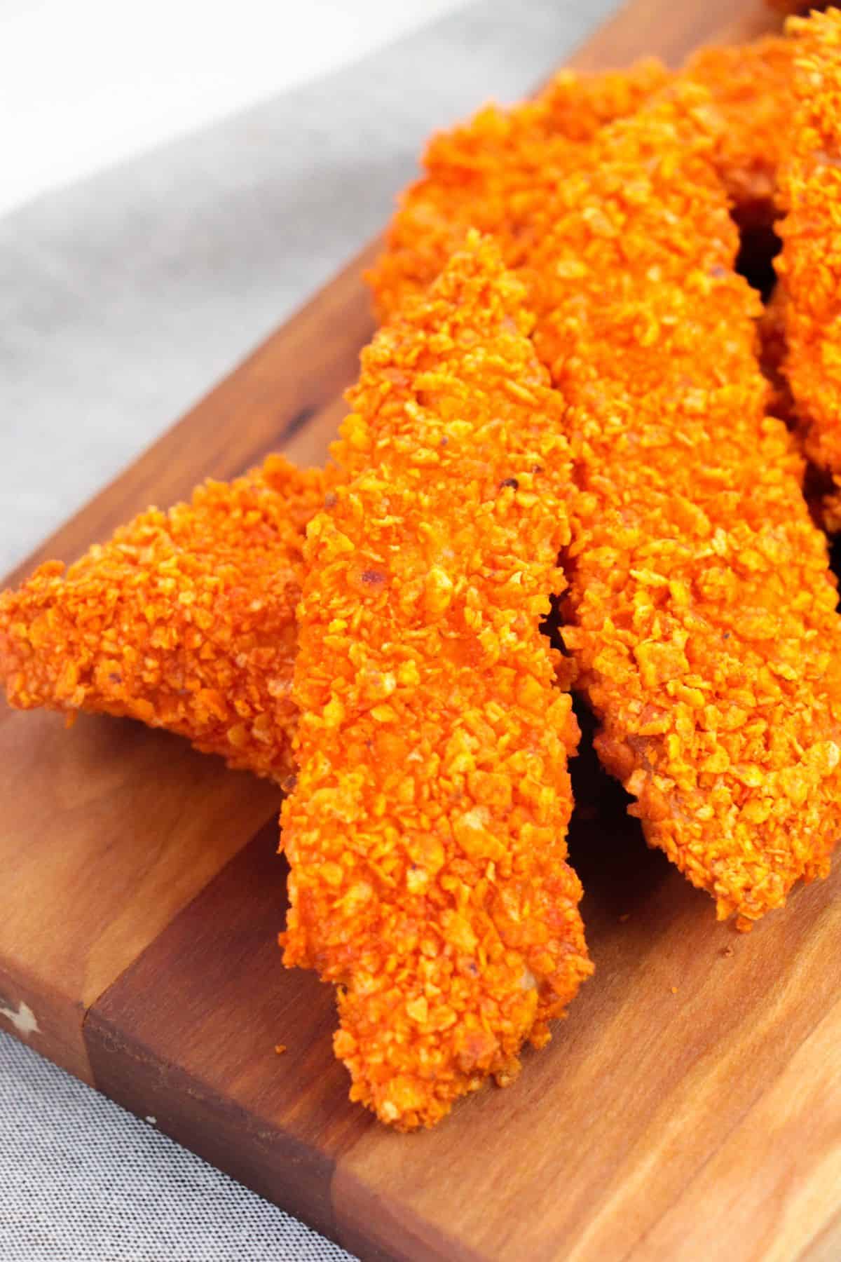 Several crispy Doritos Crusted Chicken tenders, coated in a bold orange crunch, are arranged on a wooden cutting board.