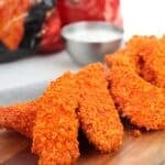 Three crispy Doritos Crusted Chicken tenders are arranged on a wooden board, served with a cup of white dipping sauce and red chip bags in the background.