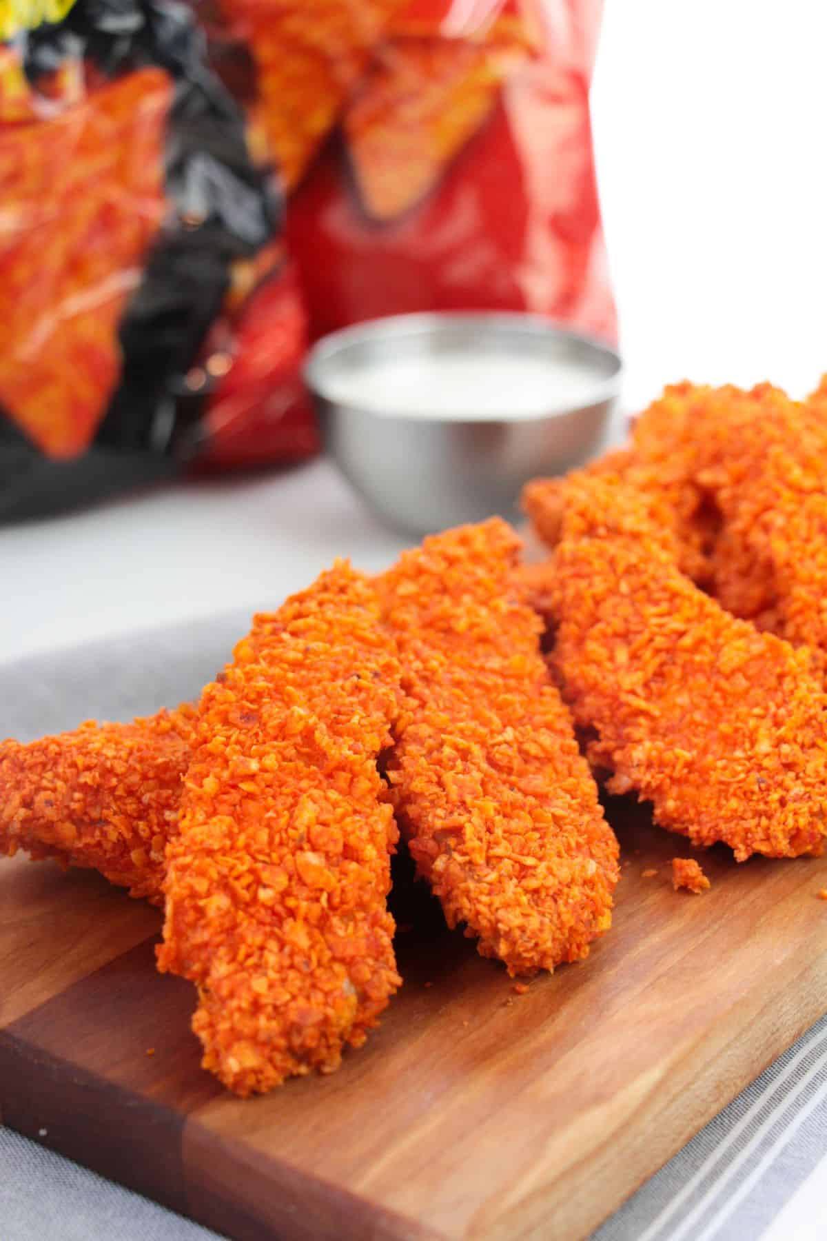 Three crispy Doritos Crusted Chicken tenders are arranged on a wooden board, served with a cup of white dipping sauce and red chip bags in the background.