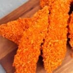 Three crispy, orange-colored Doritos Crusted Chicken tenders are arranged on a wooden cutting board.