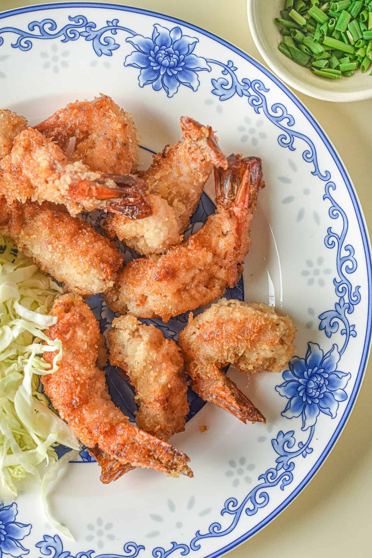 A plate of crispy panko shrimp with sliced cabbage on a blue and white floral dish, served alongside a small bowl of chopped green onions.