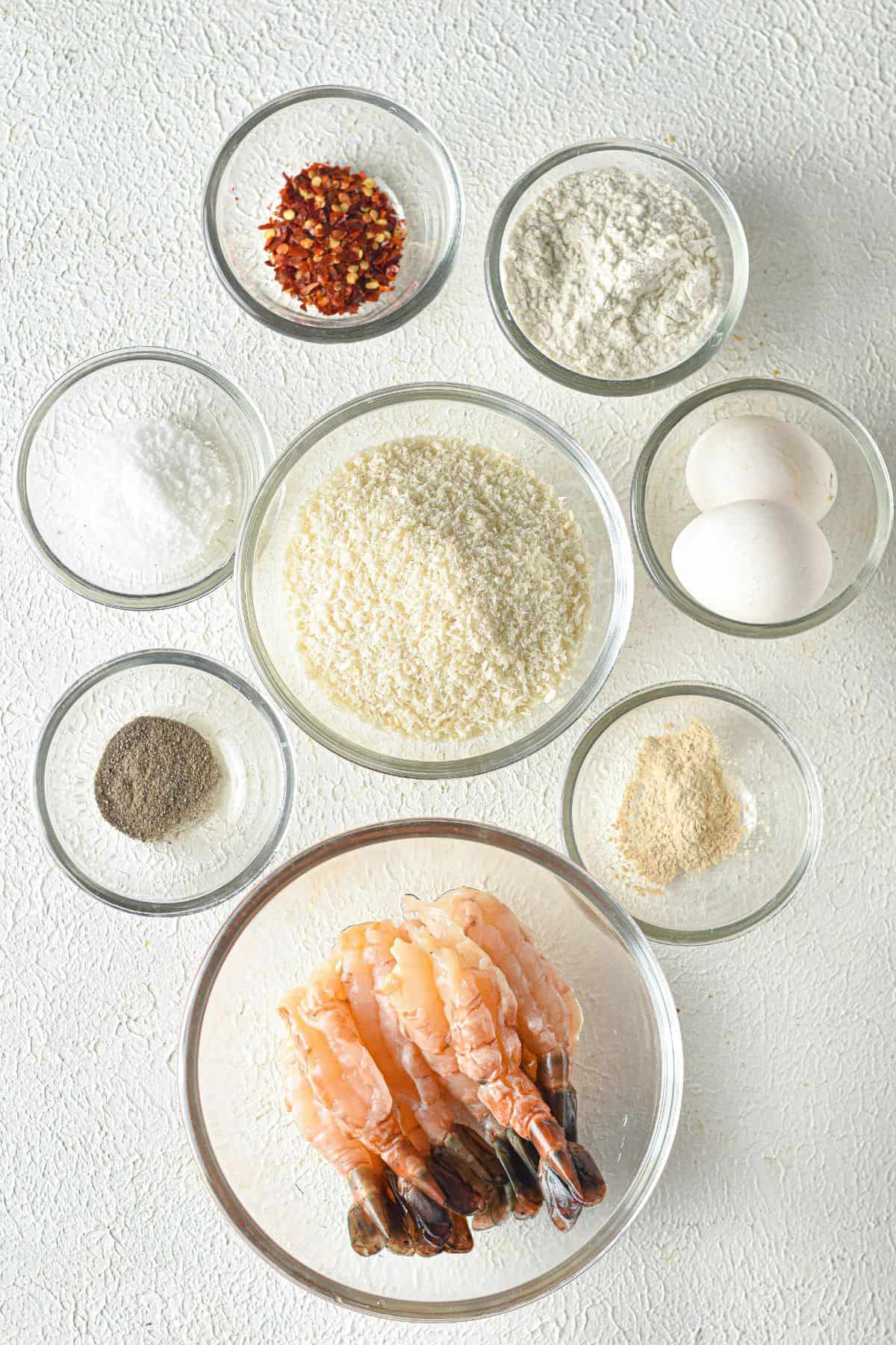 Eight glass bowls containing ingredients for Panko Shrimp—raw shrimp, panko breadcrumbs, eggs, flour, black pepper, salt, garlic powder, and red pepper flakes—are neatly arranged on a white surface.
