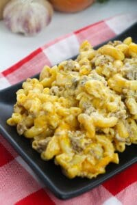 A black plate with Homemade Cheeseburger Macaroni—macaroni and cheese mixed with ground beef—sits on a red and white checkered cloth. Garlic and onion are in the background.