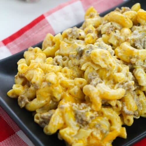 A black plate with Homemade Cheeseburger Macaroni—macaroni and cheese mixed with ground beef—sits on a red and white checkered cloth. Garlic and onion are in the background.