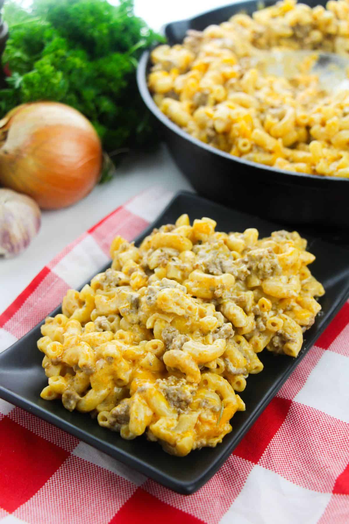 A black plate with Homemade Cheeseburger Macaroni—macaroni and cheese mixed with ground meat—placed on a red and white checkered cloth; skillet of the same dish and vegetables in the background.