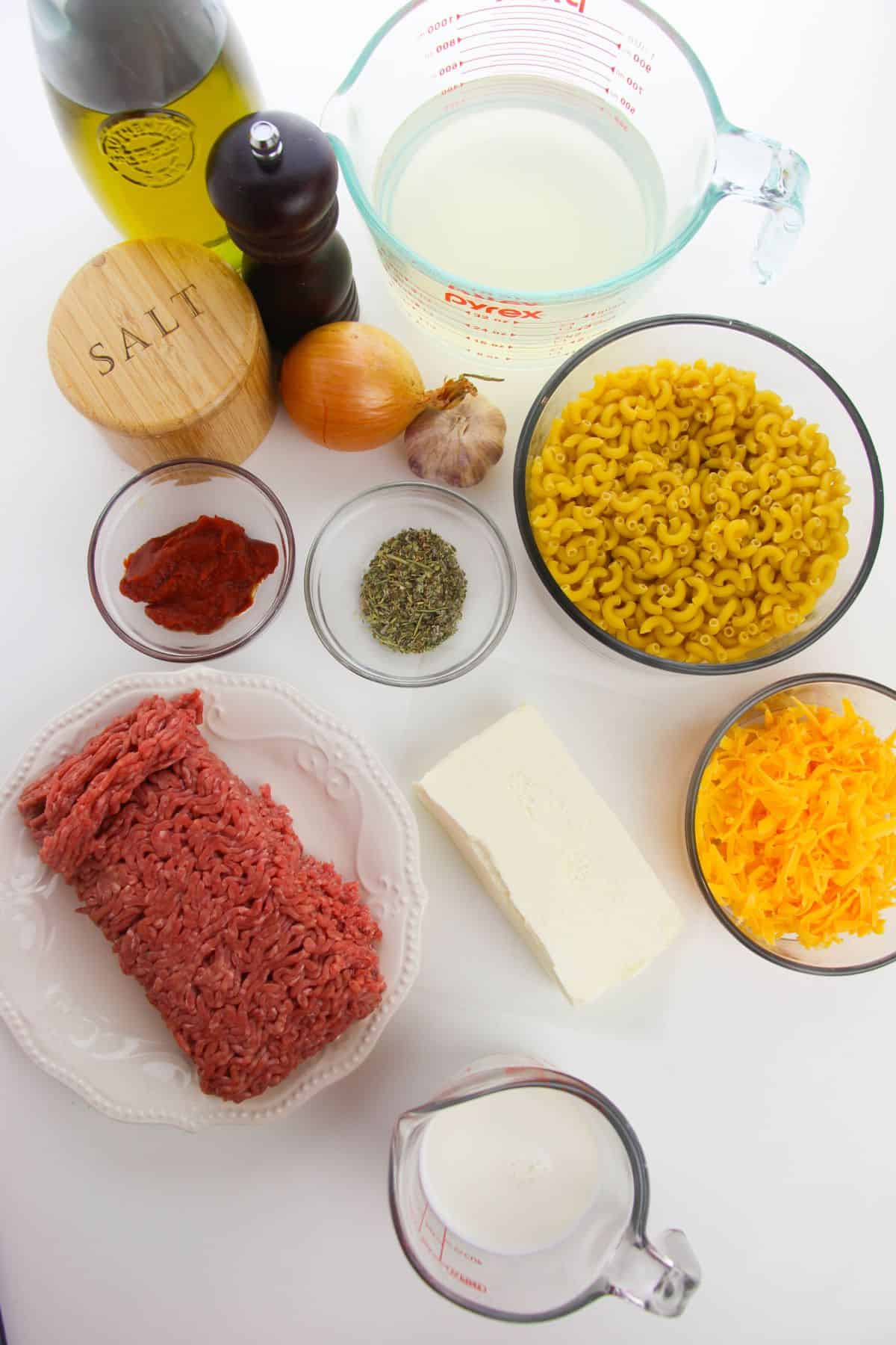 Ingredients for Homemade Cheeseburger Macaroni arranged on a white surface, including ground beef, elbow macaroni, cheese, onion, garlic, tomato paste, seasonings, butter, milk, broth, oil, salt, and pepper.