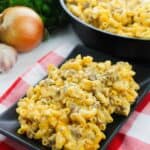 A serving of Homemade Cheeseburger Macaroni with ground meat and cheese sits on a black plate, while a skillet of the same dish and fresh vegetables add color in the background.