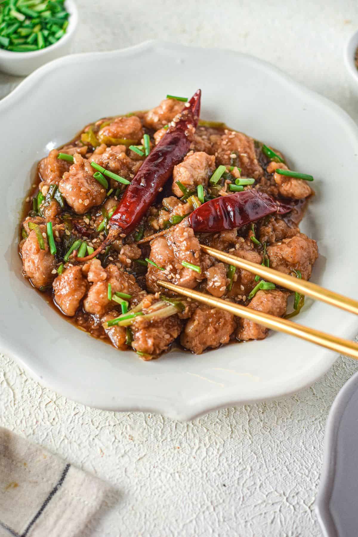 A plate of stir-fried meat, inspired by a Mongolian Chicken Recipe, garnished with dried red chilies and chopped green onions, with chopsticks resting on the side.