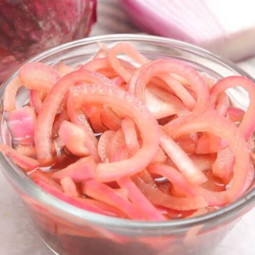 A clear glass bowl filled with Quick Pickled Onions, with a red onion and fresh onion slices in the background.