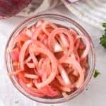 A glass bowl filled with Quick Pickled Onions—tangy pink slices—sits alongside a red onion, striped cloth, and parsley on a white surface.