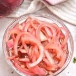 A glass bowl filled with Quick Pickled Onions sits on a white surface next to a halved red onion and a striped cloth.