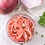 A bowl of Quick Pickled Onions sits on a white surface, alongside a whole red onion, a halved red onion, and fresh parsley.