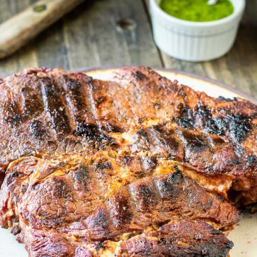 A large grilled chuck roast sits on a cutting board next to some green sauce