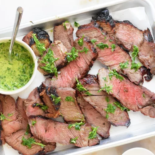 A plate is loaded with slices of grilled chuck steak with a small bowl of green sauce