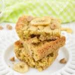 Two Peanut Butter Banana Bars with banana chips and caramel bits are stacked on a white plate, with a green checkered cloth in the background.