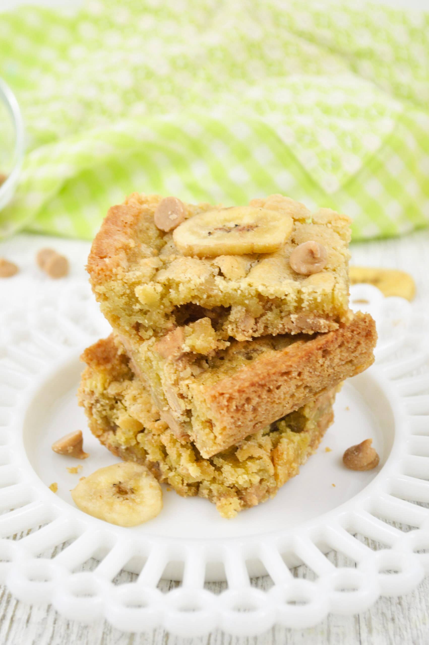 Two Peanut Butter Banana Bars with banana chips and caramel bits are stacked on a white plate, with a green checkered cloth in the background.