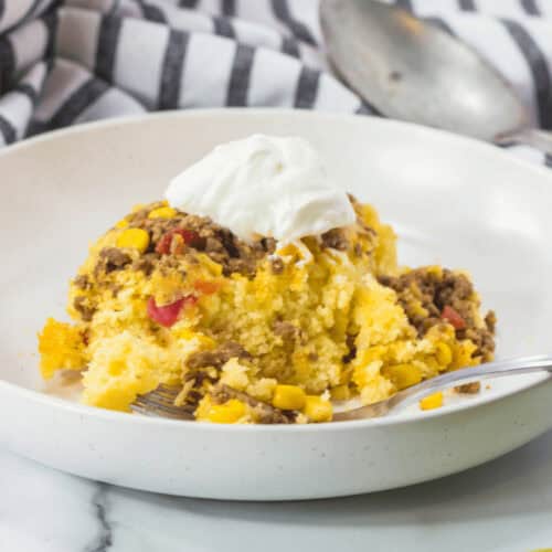 A bowl of Cowboy Cornbread Casserole with mixed vegetables, ground meat, and a dollop of sour cream, accompanied by a spoon on a striped cloth background.