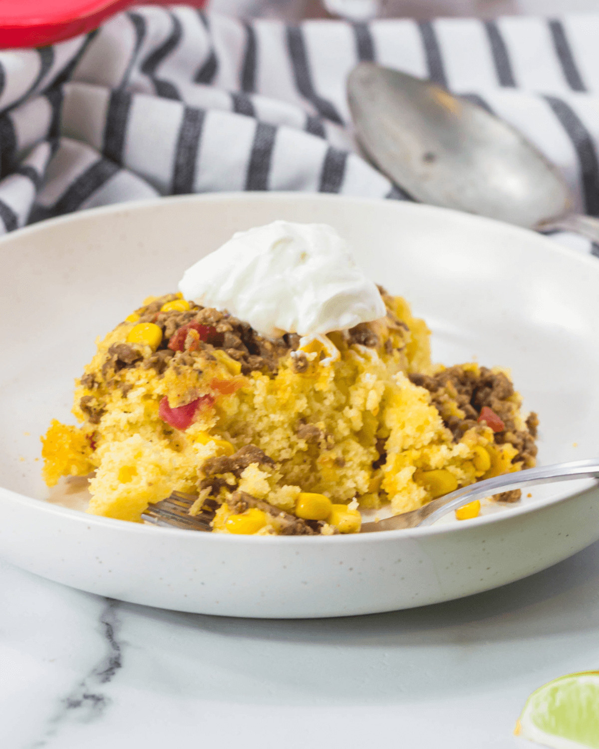 A bowl of Cowboy Cornbread Casserole with mixed vegetables, ground meat, and a dollop of sour cream, accompanied by a spoon on a striped cloth background.