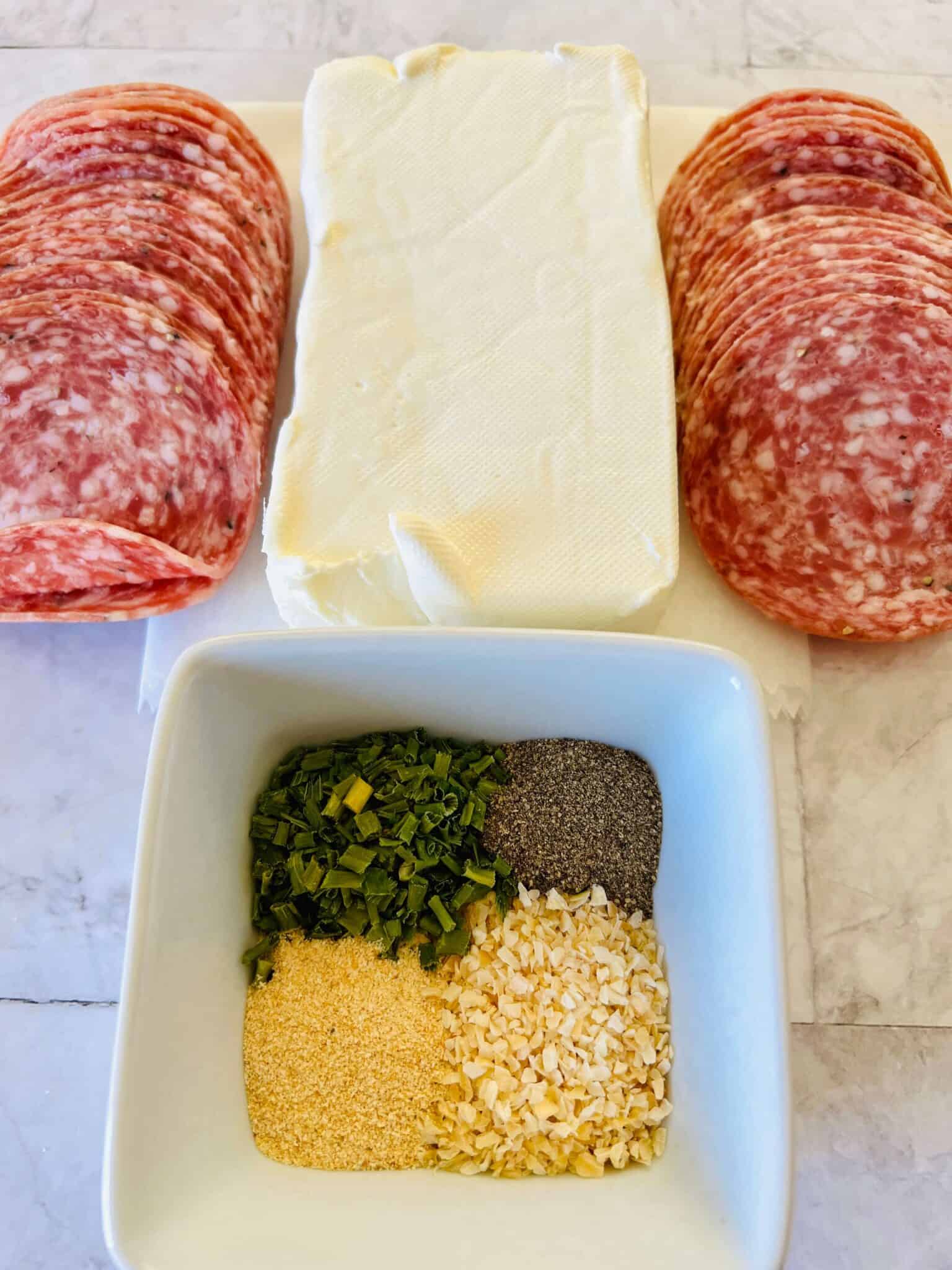 Salami roll ups are made with salami slices and a block of cream cheese, perfectly complemented by a square bowl filled with chopped herbs, black pepper, garlic powder, and dried minced onion.
