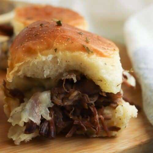 A Beef Sliders sandwich with shredded beef and melted cheese rests on a soft bun atop a wooden board, while another slider and a white cloth appear in the background.
