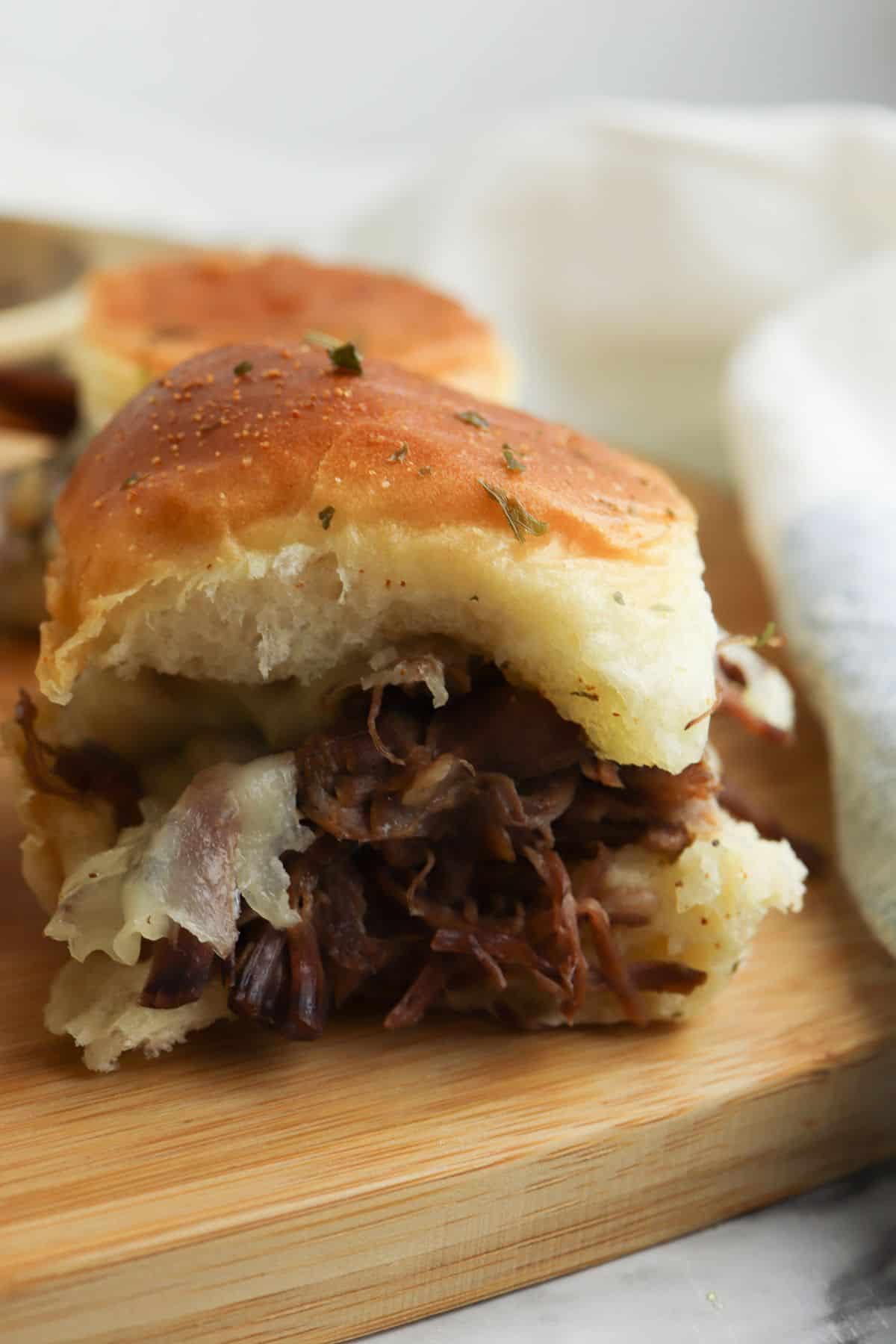 A Beef Sliders sandwich with shredded beef and melted cheese rests on a soft bun atop a wooden board, while another slider and a white cloth appear in the background.