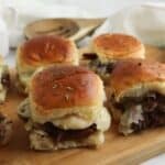 Sandwiches with melted cheese on golden buns are arranged on a wooden cutting board, with a white napkin and wooden utensils in the background.