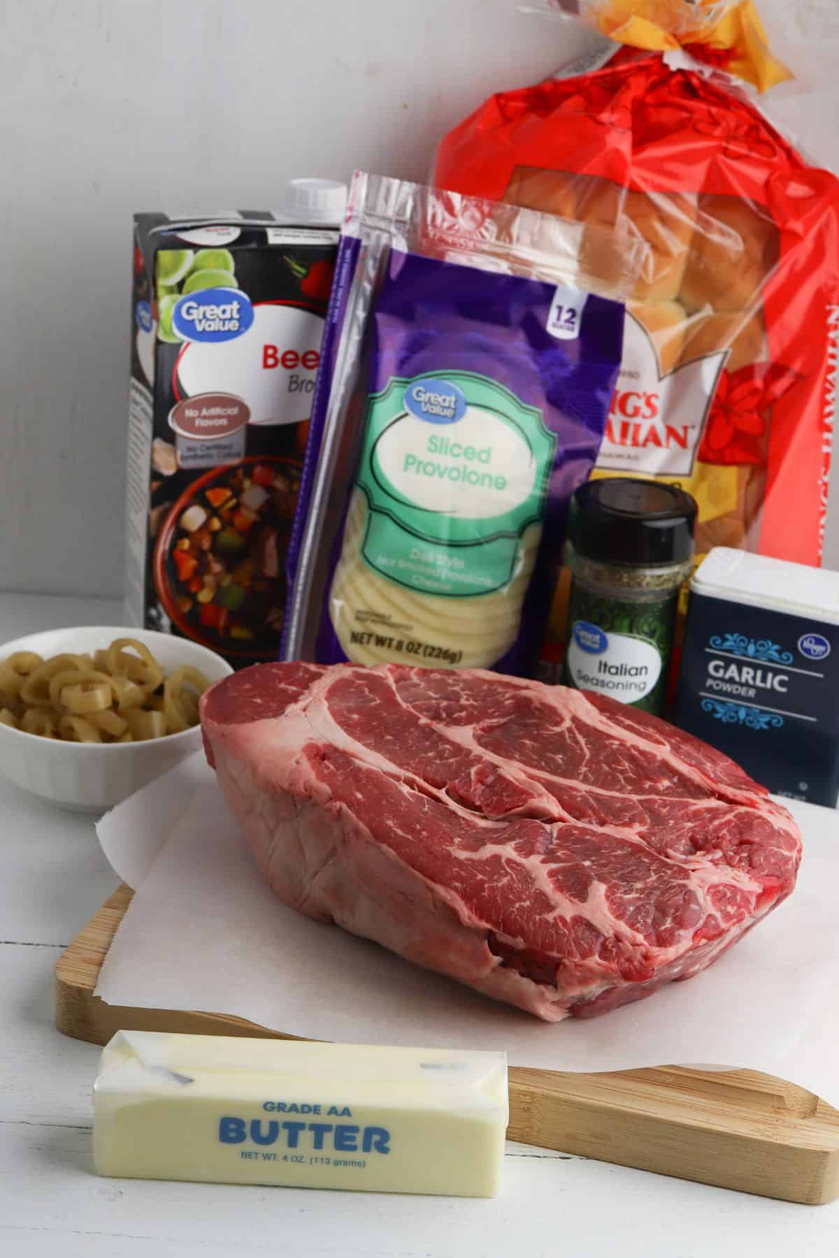 Raw chuck roast on parchment with a stick of butter, sliced provolone cheese, Italian seasoning, jarred garlic, pepperoncini, beef broth, and a loaf of Italian bread.