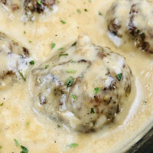 Close-up of Easy Swedish Meatballs Recipe: tender meatballs covered in creamy sauce, garnished with chopped herbs, in a black skillet.