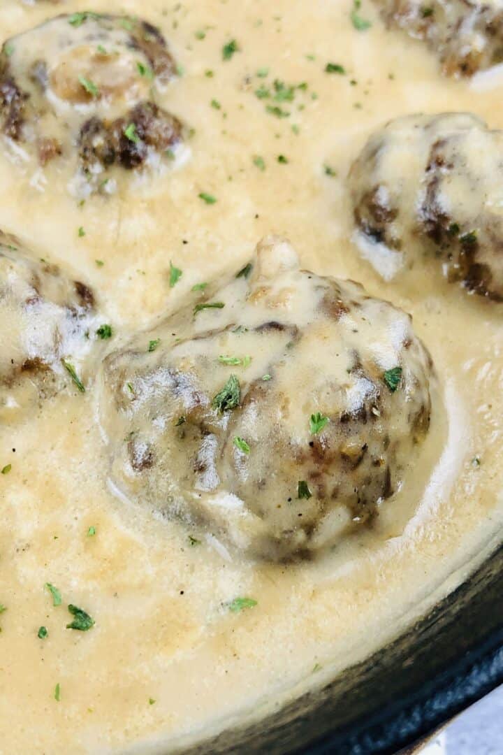 Close-up of Easy Swedish Meatballs Recipe: tender meatballs covered in creamy sauce, garnished with chopped herbs, in a black skillet.