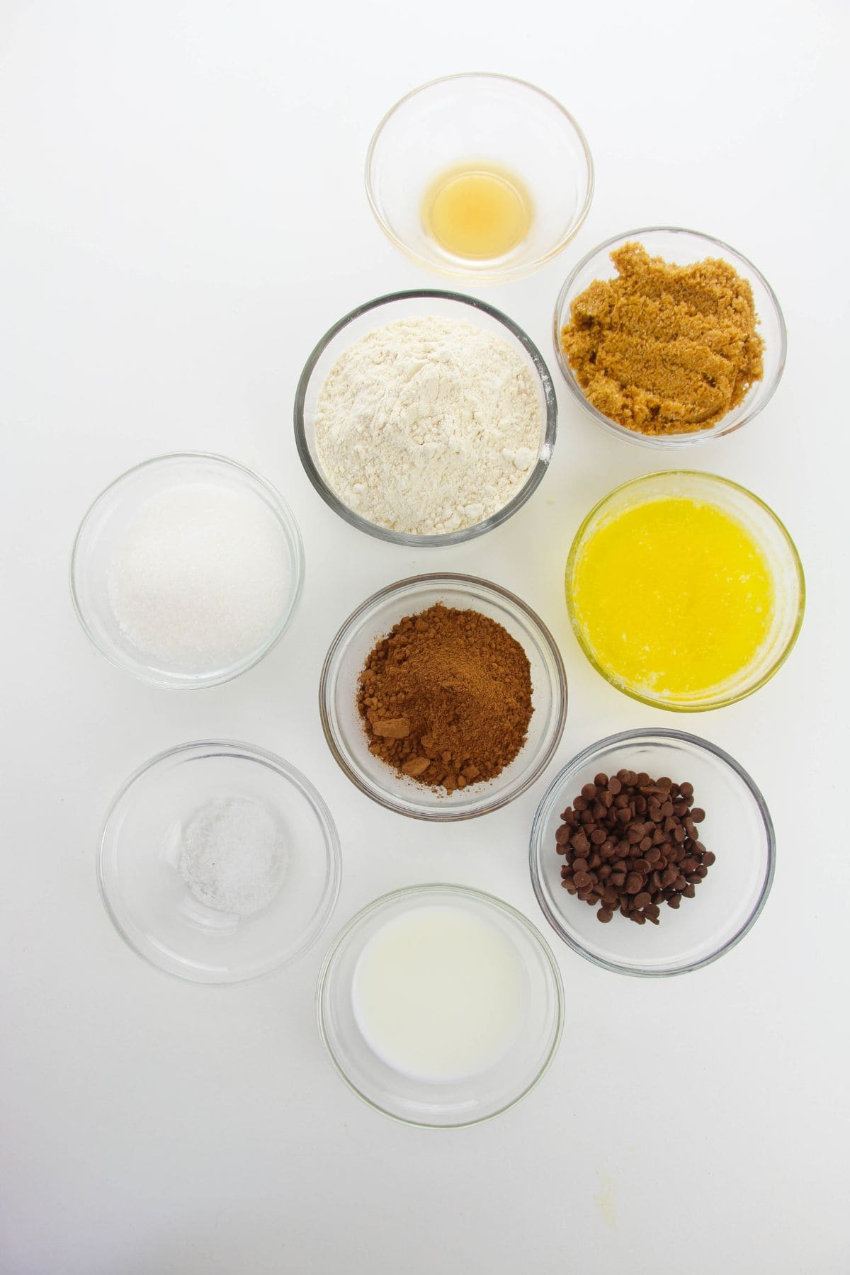 Nine glass bowls filled with various baking ingredients, including flour, brown sugar, white sugar, cocoa powder, melted butter, chocolate chips, vanilla extract, milk, and salt on a white background.