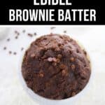 A bowl of Edible Brownie Batter topped with chocolate chips sits on a white surface, with more chocolate chips scattered in the background.