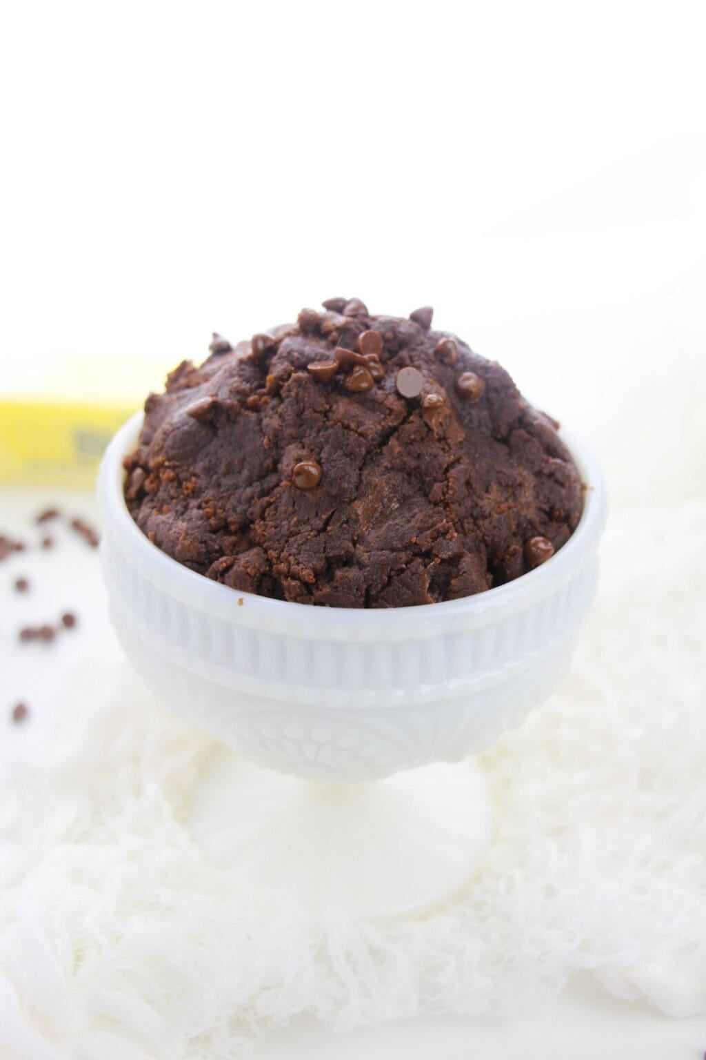 A white pedestal bowl filled with rich edible brownie batter topped with chocolate chips, set on a white textured surface.