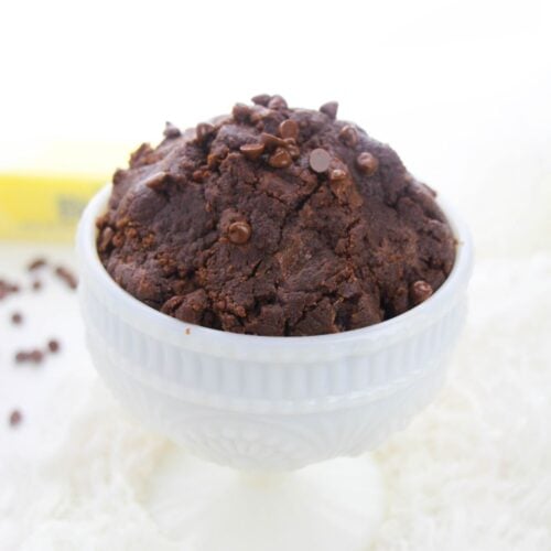 A white pedestal bowl filled with rich edible brownie batter topped with chocolate chips, set on a white textured surface.