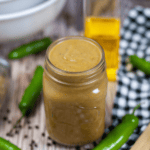 A jar of homemade green chili enchilada sauce on a wooden table, surrounded by fresh jalapeños, black pepper, and a bottle of oil.